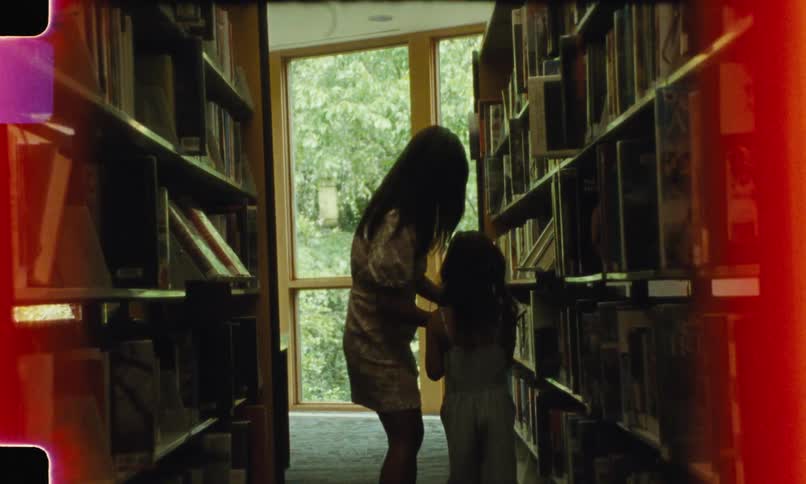 A mother helps her daughter take a book from a library shelf