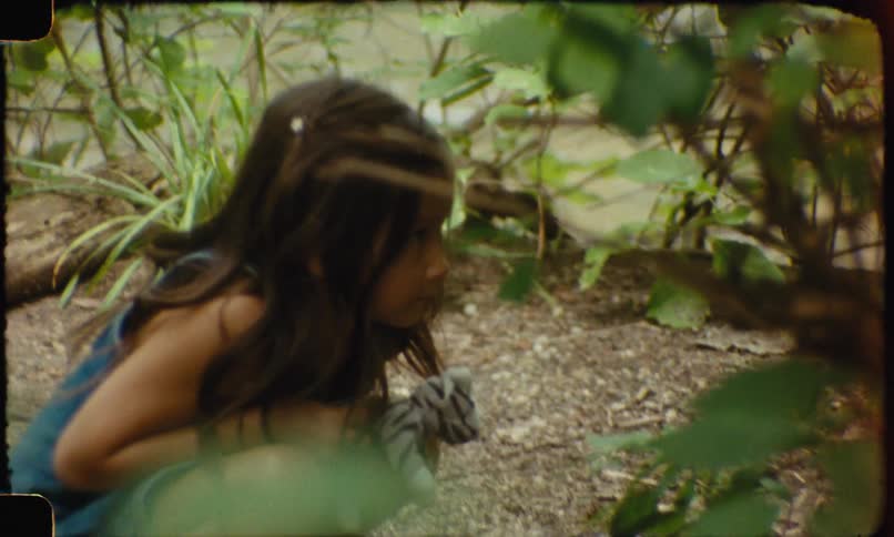 A young girl in a blue denim outfit crouches in a wooded area appearing to play or explore among the trees and undergrowth