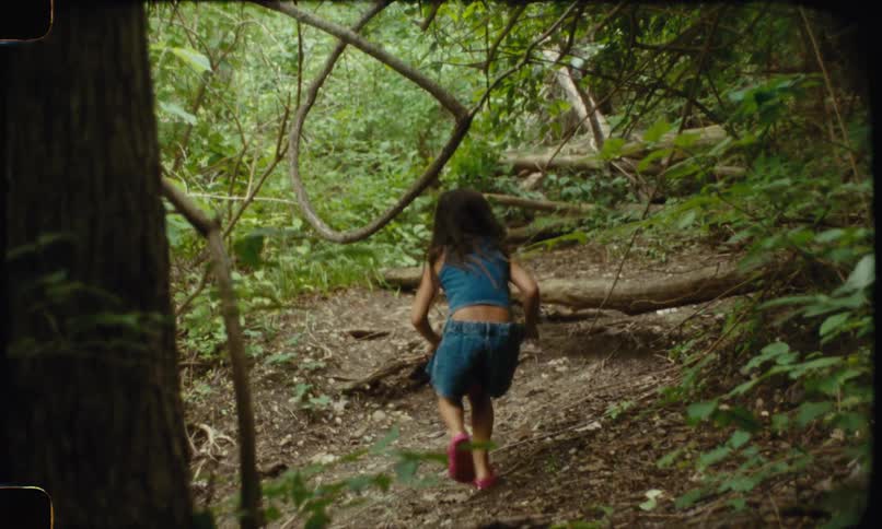 A little girl with long dark hair wearing a sleeveless denim top and denim shorts is walking up a dirt and tree covered hill in a dark forest playing hide and seek