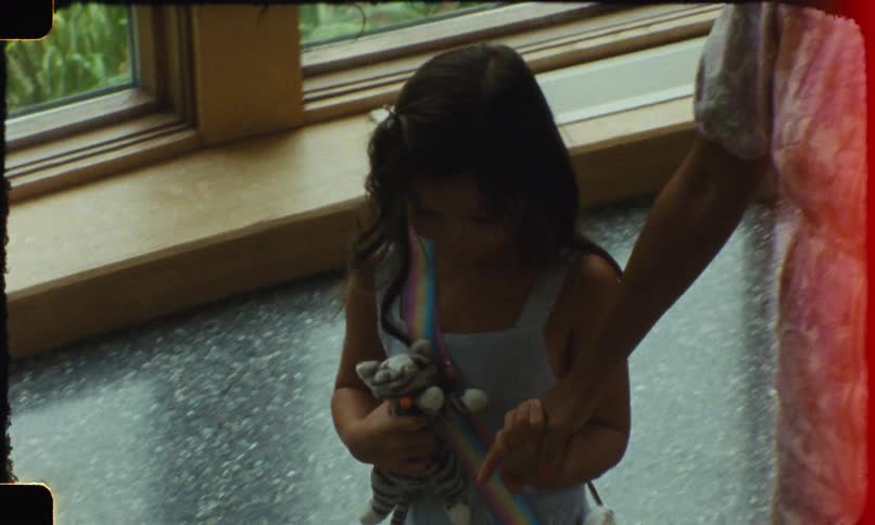 A little girl with long dark hair wearing a light colored dress and holding a toy is standing on a staircase holding the hand of an adult woman as they walk down the stairs