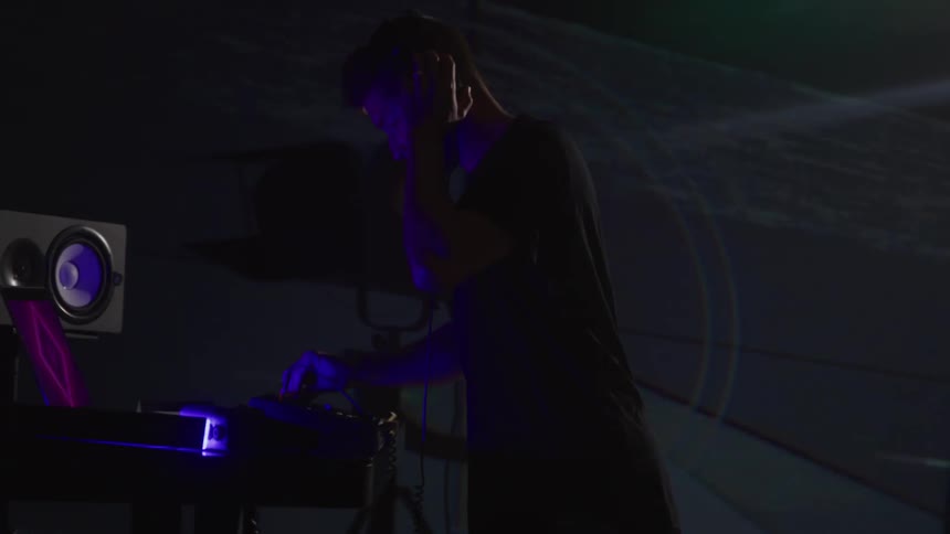 A dj wearing a dark t shirt and a backwards baseball cap is standing in a dimly lit studio wearing headphones as they use a turntable and a mixer while a laptop and a large speaker