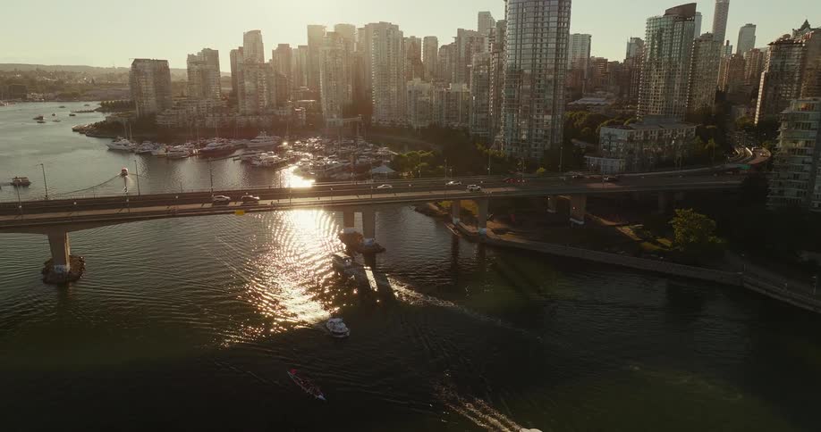 An aerial view shows a citys skyline with a bridge and a marina below as the sun sets with a handful of boats in the water and cars crossing the bridge