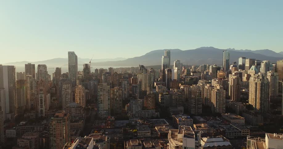 The video is a wide elevated shot of a large city with a mountain range in the distance the buildings are a mix of older and modern highrises and the sun is low in the sky casting a warm golden light over the urban landscape
