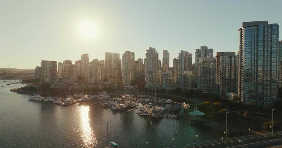 An aerial view shows a large city with numerous tall buildings and a harbor full of boats and yachts with a bridge running horizontally in the foreground and a bright sun in the sky reflecting on the water