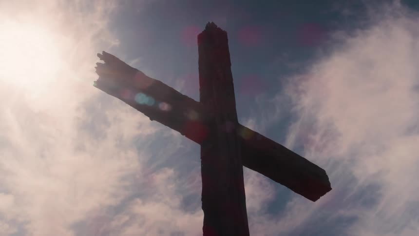 A lowangle shot of a large weathered wooden cross is silhouetted against a bright sky with fastmoving clouds and a strong sun flare