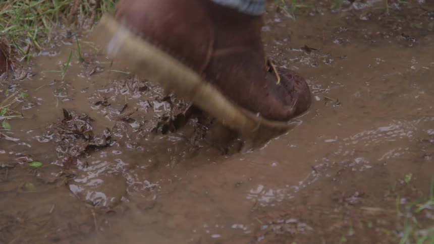 A person wearing blue jeans and a brown leather boot with yellow laces steps into a muddy puddle causing brown water and mud to splash up around the foot