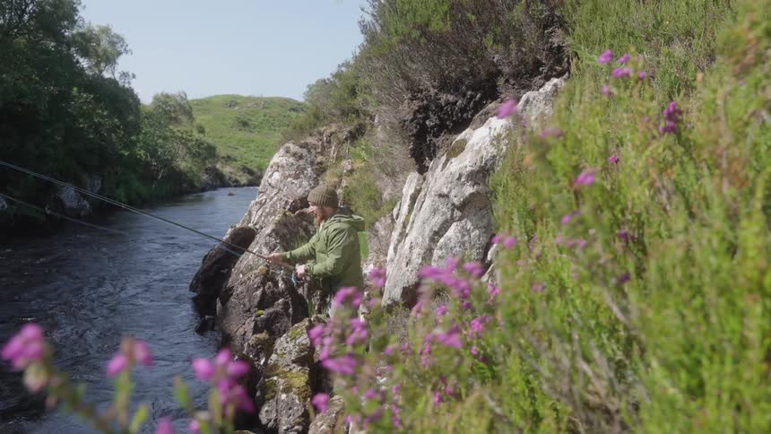 Man fishing in a rocky river surrounded by vegetation and purple flowers