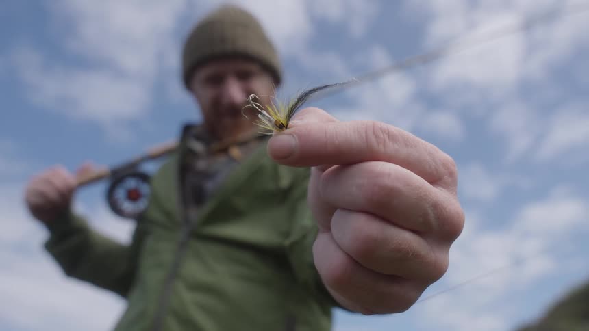 Based on the video a man is shown outdoors holding a fly fishing lure close to the camera he is wearing a knit hat and a jacket and a fishing rod is visible over his shoulder