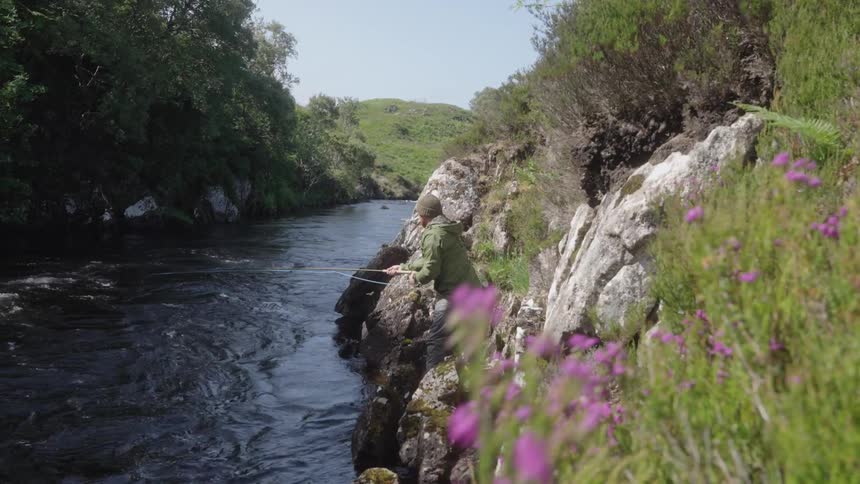 A person fishes while standing on rocky banks beside a flowing river under a sunny sky