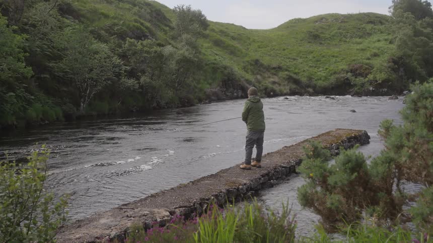 A person wearing a dark green jacket and brown boots is standing on a narrow stone walkway in the middle of a flowing river casting a fishing line with lush green hills and trees lining both sides of the riverbank