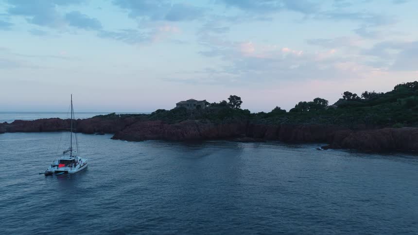 A catamaran yacht floats near a rocky coastal cliff at dusk