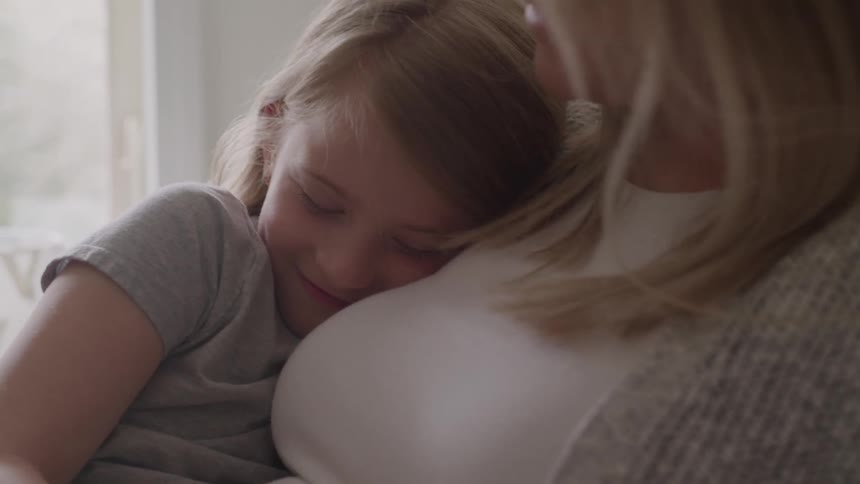 A little girl with light colored hair is resting her head on a womans shoulder with her eyes closed and a calm expression on her face while the woman looks down at her with a small smile