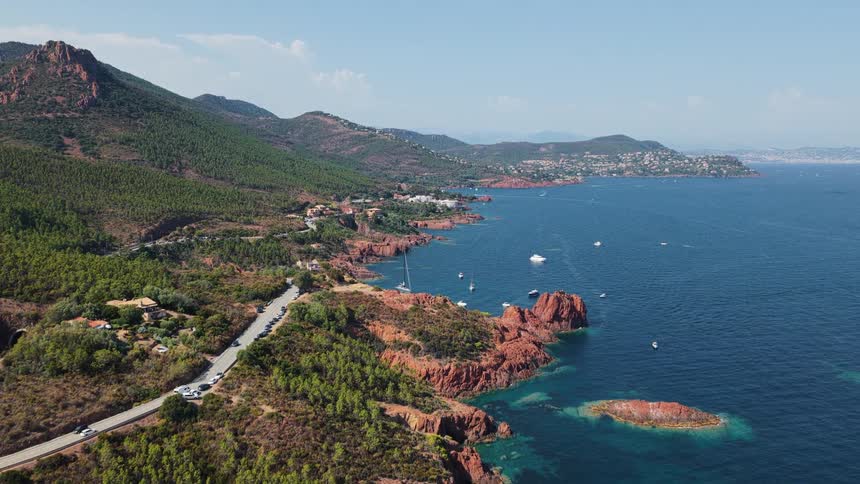 An aerial view of a vibrant blue ocean shows a rocky coastline with red cliffs and a winding road along the shoreline with several boats anchored in the water and a town visible in the distance
