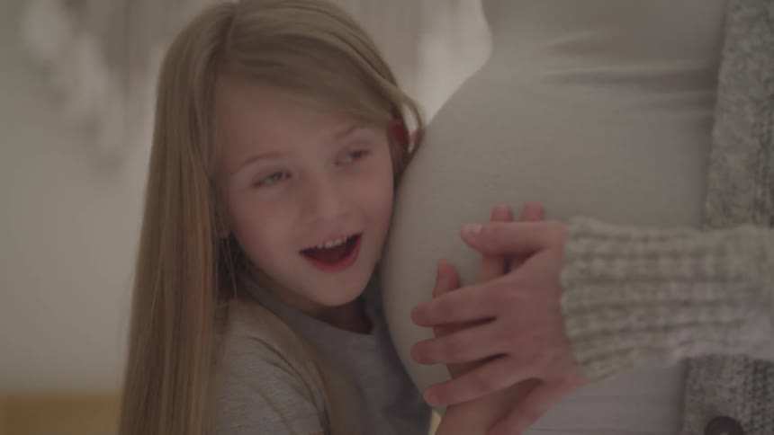 A little girl hugs her mothers pregnant belly with a shy smile