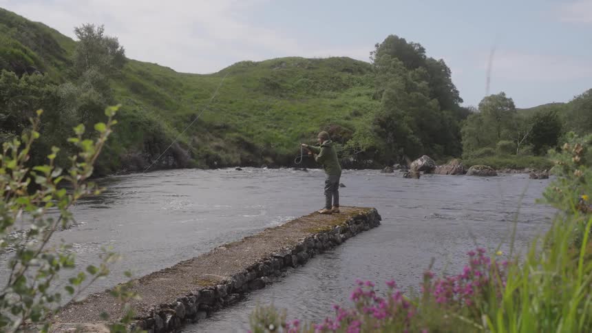 Person fishes alone in a river surrounded by green hills