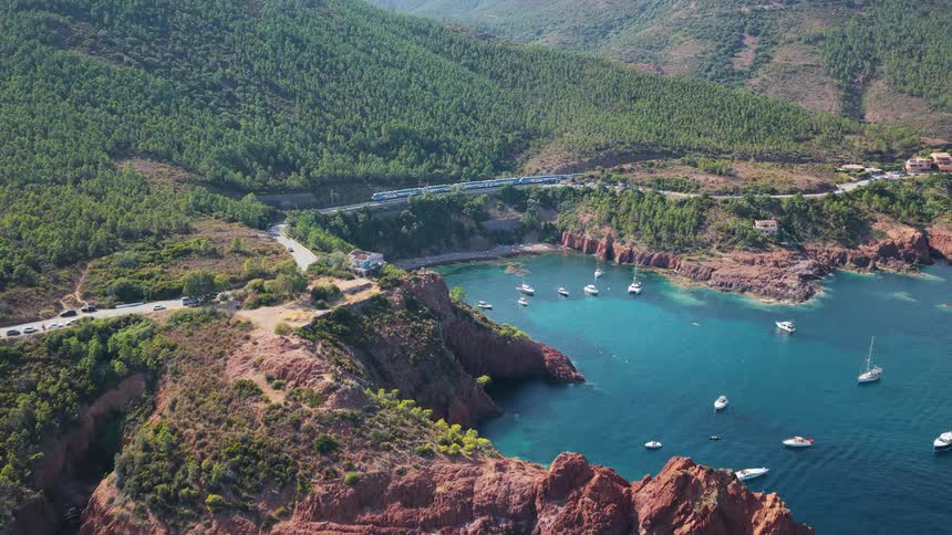 An aerial view shows a blue and white train traveling along a railway track that runs through a green wooded mountain range near a red rocky coastline with a tranquil blue cove full of anchored boats