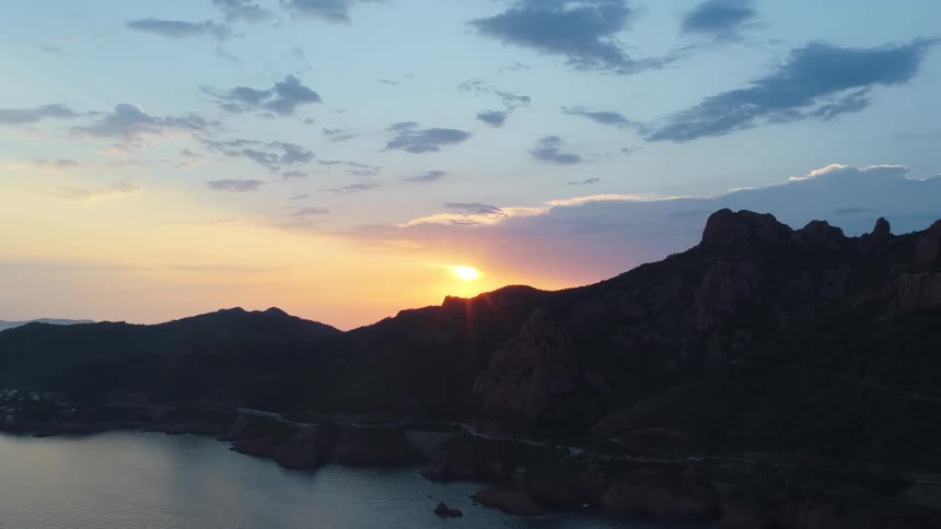 An aerial view of a calm body of water shows a rugged rocky mountain range silhouetted against a cloudy sky with the setting sun visible behind it with small houses visible along the shoreline