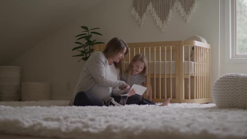 Pregnant woman sitting in the room shows pictures to her daughter