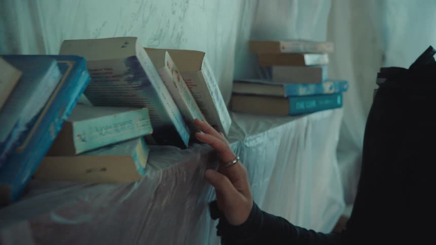 A close view of a persons hand with a silver ring on the index finger shows it resting on the edge of a table covered by a semi opaque plastic sheet with several books stacked on top