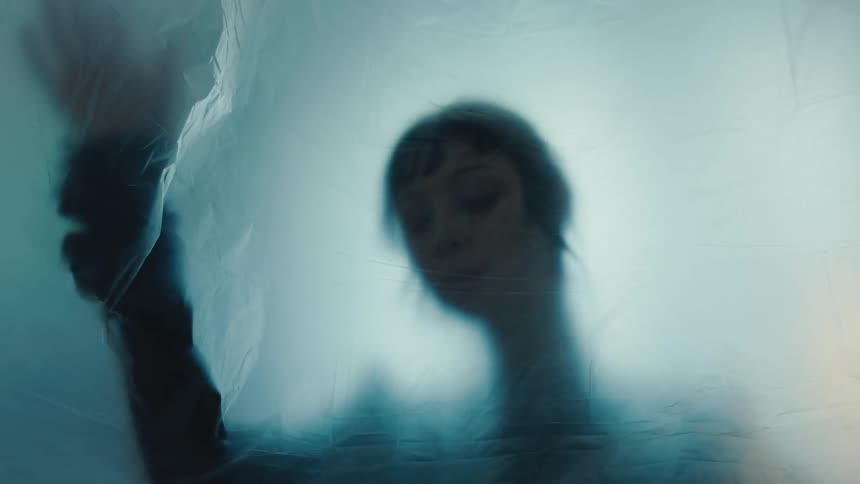 A shadowy silhouette of a woman is visible behind a light blue plastic sheet moving up and down her arms behind the sheet