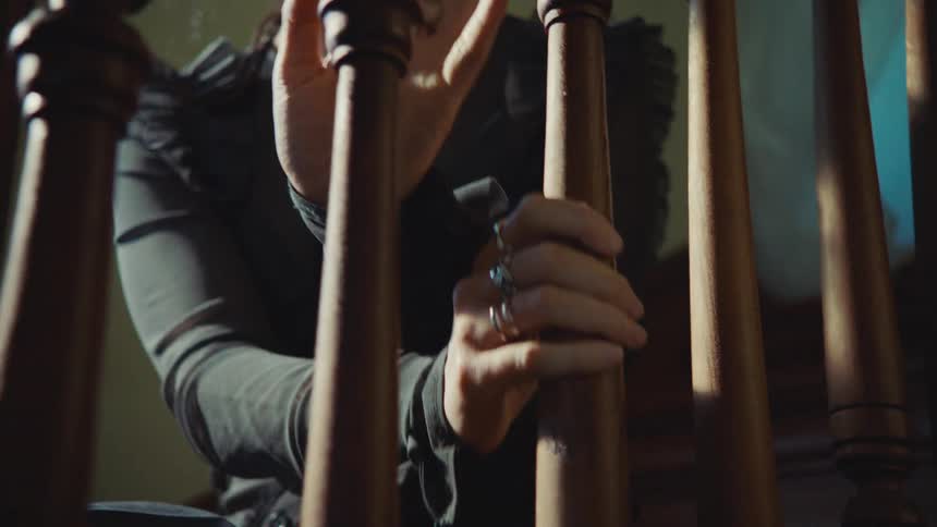 A woman wearing a dark long sleeved shirt with ruffled shoulders and several silver rings on her fingers is descending a staircase with her hands gripping the wooden balusters