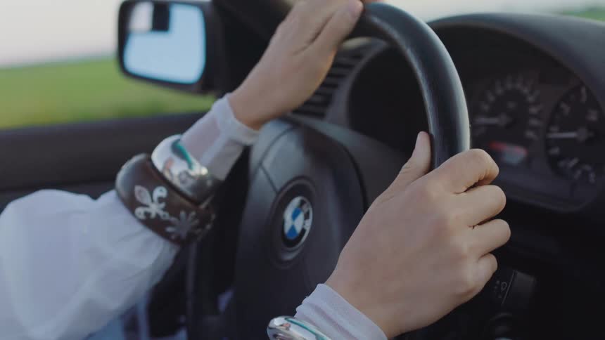 Based on the images from the video a closeup shot shows a persons hands on a steering wheel with one hand wearing a large silver bracelet the person is driving a car on a sunny day