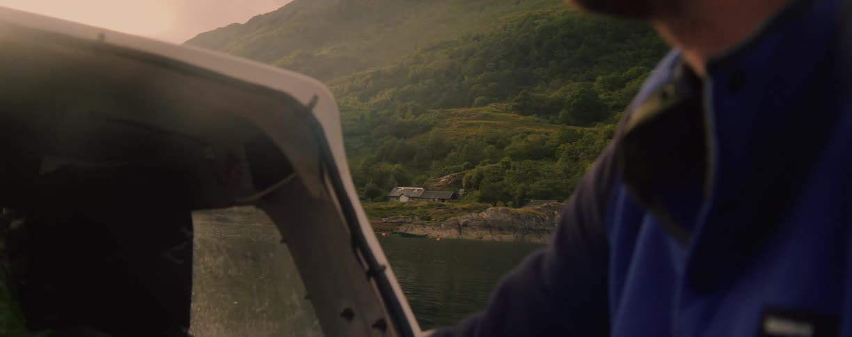 A man steers a small boat across a lake with mountains in the background
