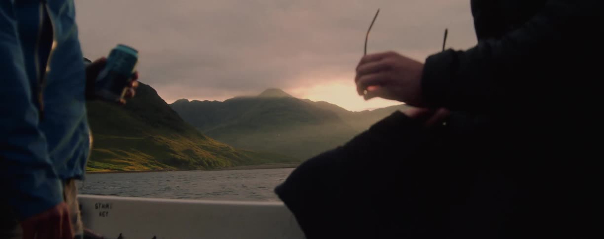 A view from a boat shows two people while moving across a calm body of water with a grassy mountain range visible in the background against a cloudy bright sunset