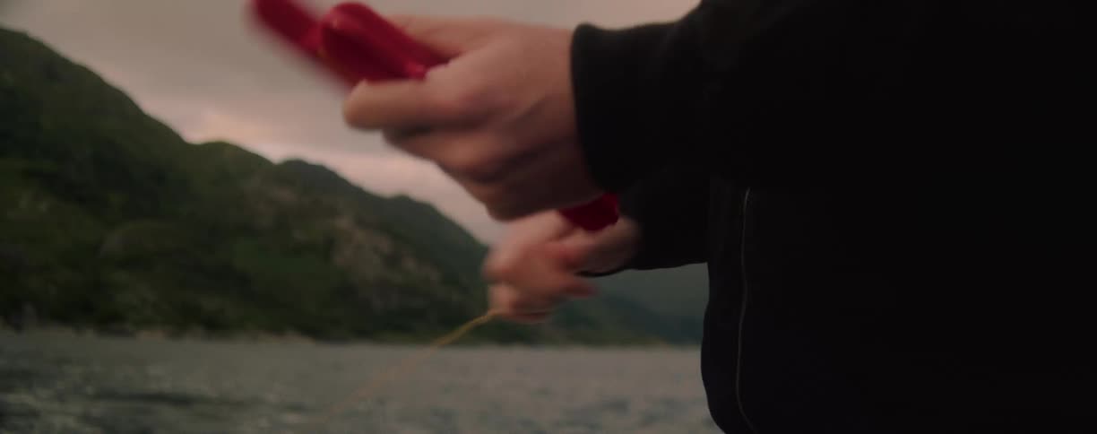 Person reels a fishing line into a small boat on a lake surrounded by mountains