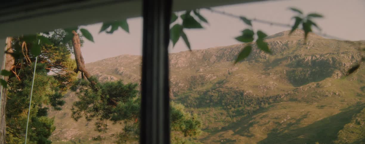View from a window reveals sunlit mountains surrounded by trees