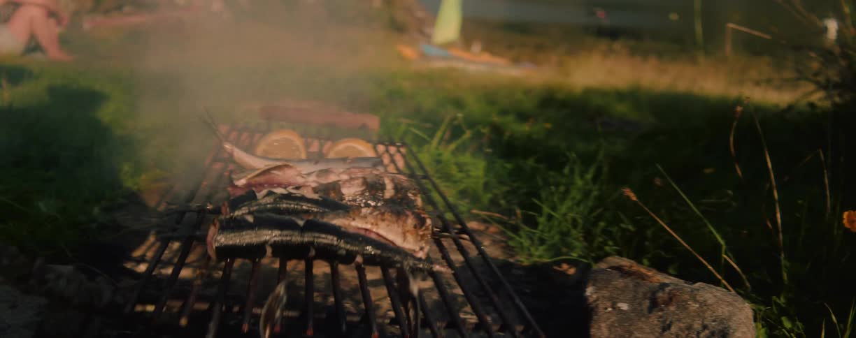 In a closeup shot whole fish garnished with lemon slices cook on a makeshift grill sending smoke into the air over a grassy outdoor area