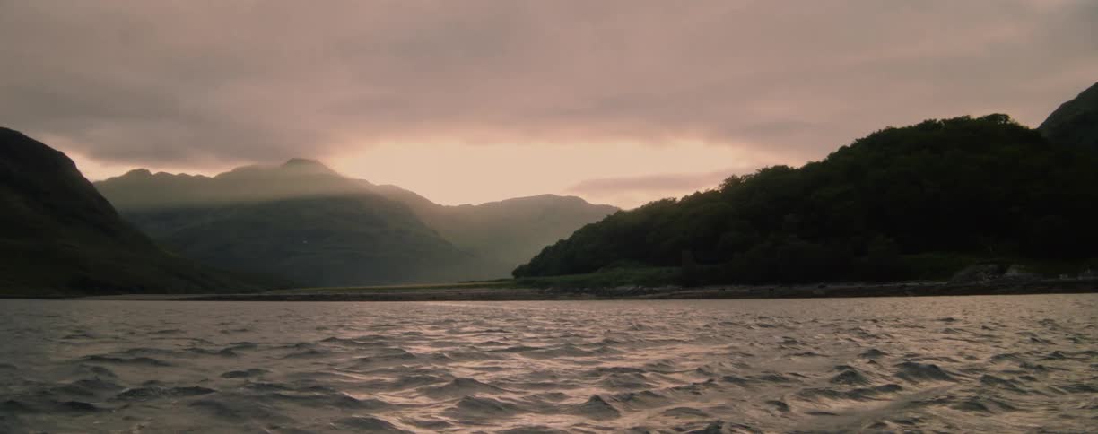 From the perspective of a boat on choppy water the camera looks out over a lake towards a dramatic mountain range at sunset under a cloudy sky