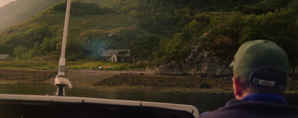A person wearing a green baseball cap is looking out from a boat toward a small white stone house on a green rocky shoreline with a few kayaks pulled onto the small beach