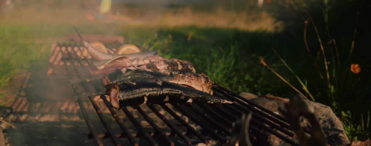 Fish are being grilled outdoors over an open flame emitting smoke with a natural grassy background