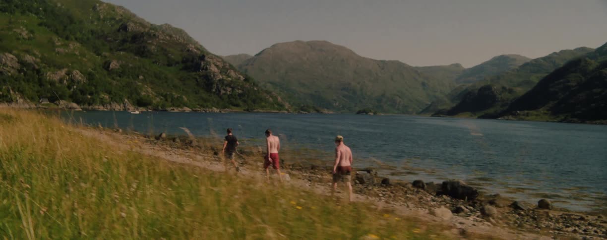Three men walk along the shore of a lake surrounded by green mountains
