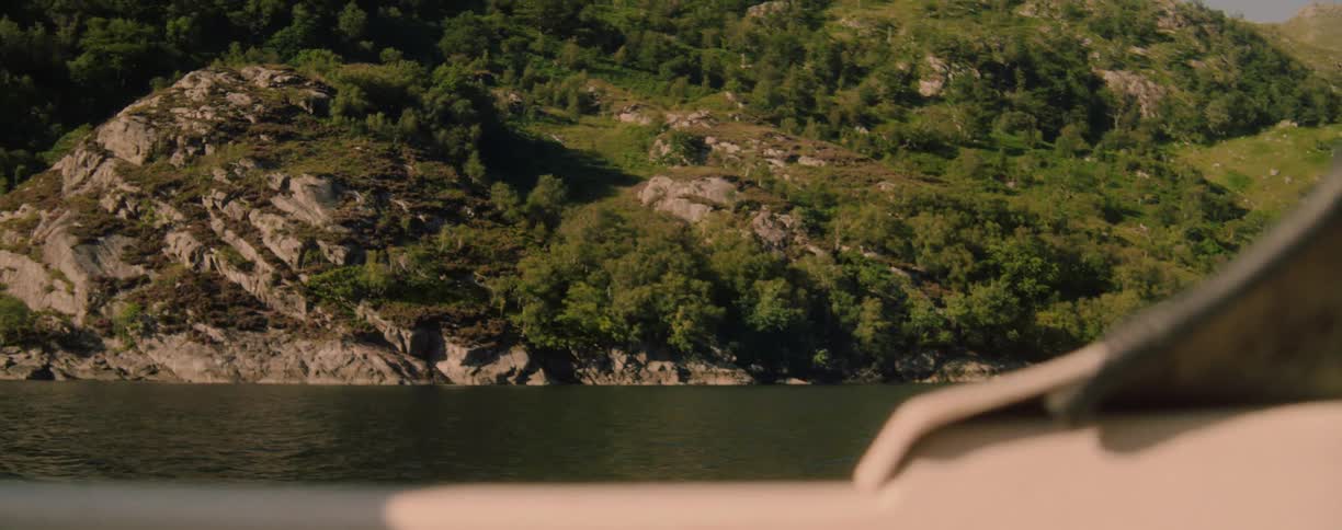 A wide view from a boat shows a large rocky hillside with dark green foliage on the slopes and trees on the summit as the boat moves across a body of water