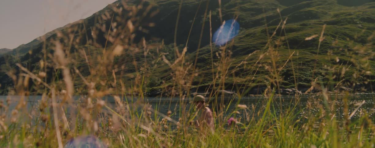 Person walking alongside tall grass in the foreground contrasts with a lake and green mountains in the background under sunlight