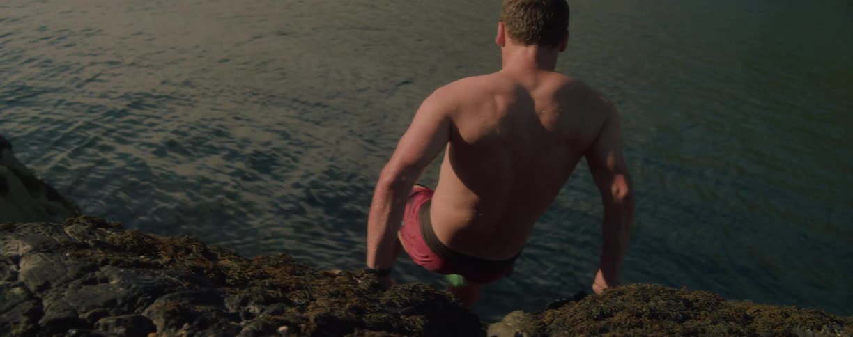 A man with his shirt off is seen from the back wearing a pair of red and black shorts as he stands on the edge of a rocky cliff to jump into the blue water below