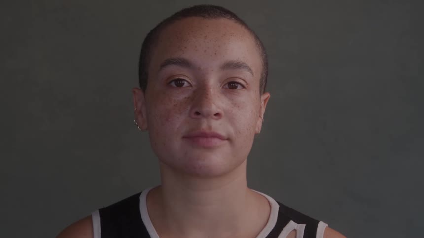 A person with very short dark hair and freckles on their face and shoulders is shown in a side view looking to the left with a neutral expression before turning to look directly at the camera