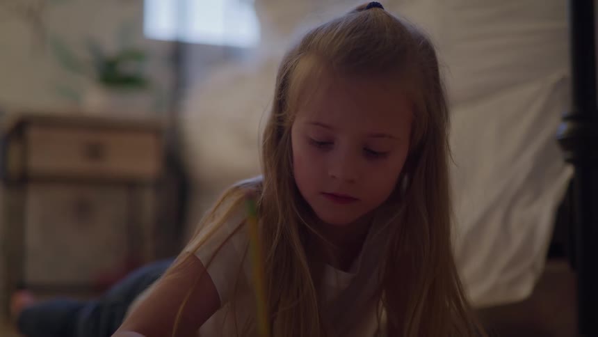 A young girl lays on her stomach while writing with a pencil