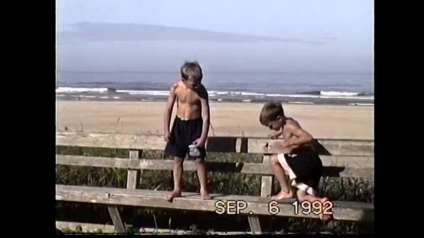 The video which has a timestamp of sep 6 1992 shows two young boys at the beach they stand on a wooden fence facing the camera flexing their muscles and smiling with the ocean in the background