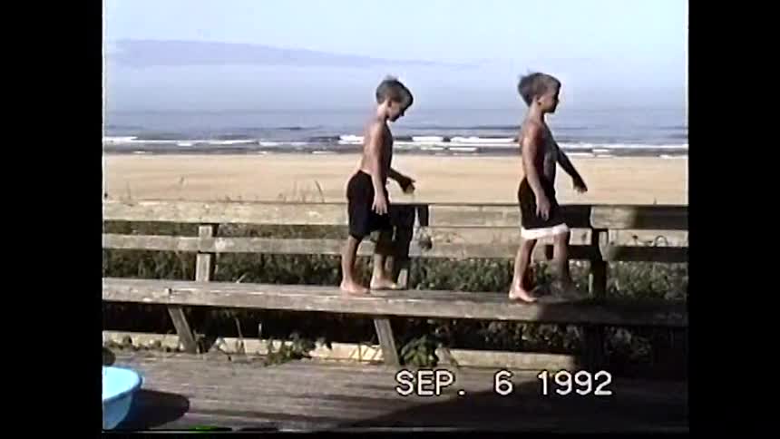 Two shirtless boys with light hair are standing on a wooden bench on a boardwalk overlooking the ocean flexing their muscles at the camera with a digital timestamp in the bottom right corner