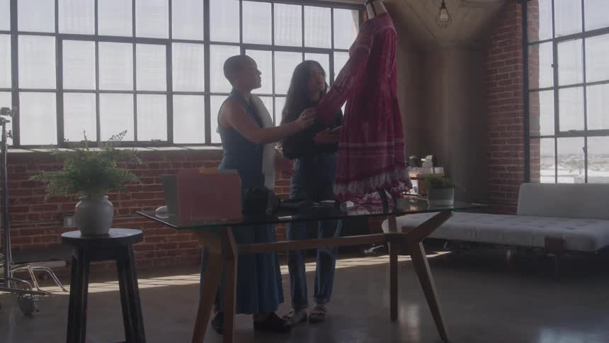 Two women standing in a welllit urban loft focusing their attention on a burgundy dress that is hanging on a table