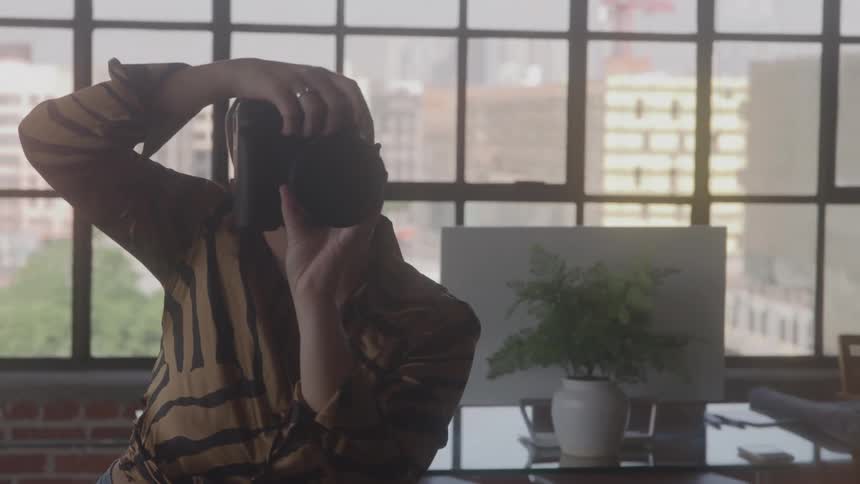 A person with a shaved head and a striped shirt is holding a camera up to their face and taking a photograph while standing in front of a large window with a plant on a desk