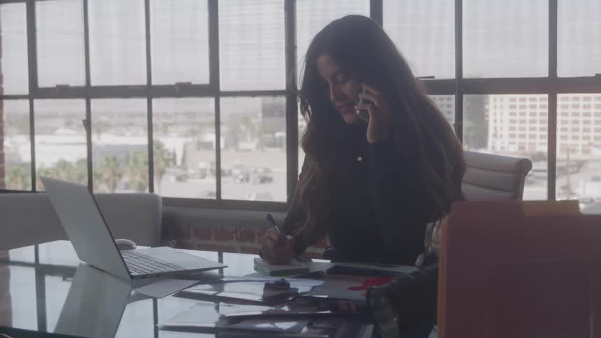 A professional woman at her office desk engaged in a phone conversation while simultaneously writing notes