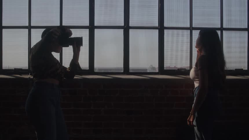 A person with dark hair is standing on the left and looking through a camera as they photograph a woman with long dark hair both standing in a dark room in front of a large sunlit window