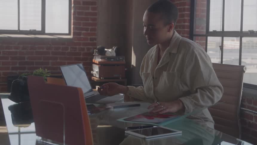 A woman with a shaved head and a neutral colored collared shirt is sitting at a large glass desk in a sunlit office with brick walls picking up and looking at documents