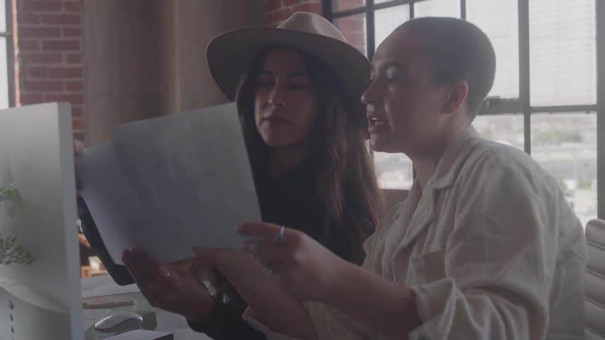 Two coworkers are seen in a brightly lit office initially working at their desks before one hands the other a paper document to review together