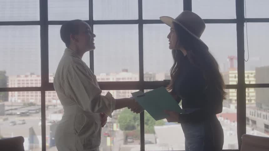 The video shows a conversation between two women standing in front of a large window with a cityscape in the background they shake hands exchange some papers and appear to be discussing business in a professional and friendly manner