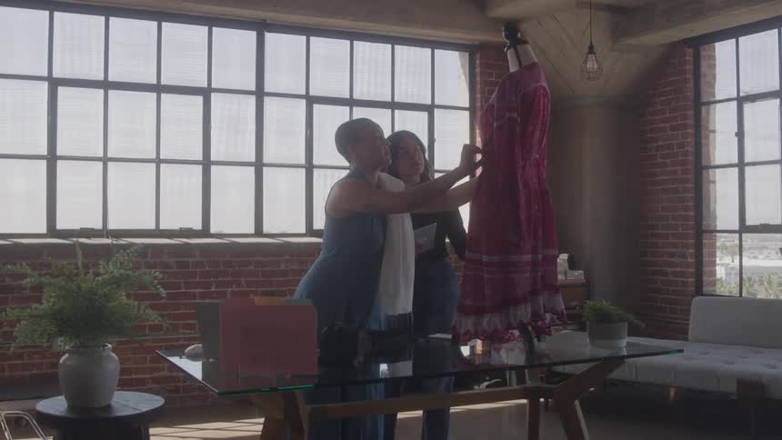Two female coworkers one with a shaved head and the other with long dark hair are standing on opposite sides of a glass desk talking as they look at a magenta and red patterned garment hanging on a mannequin